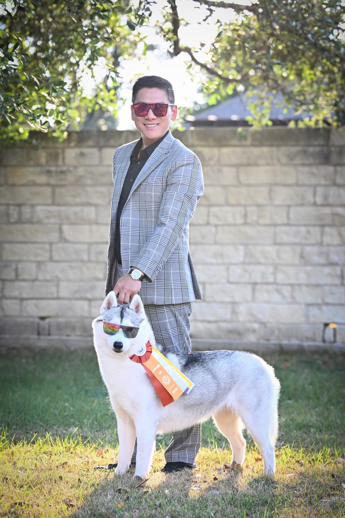 Tiny wearing sunglasses with ribbons beside her handler in a gray plaid suit