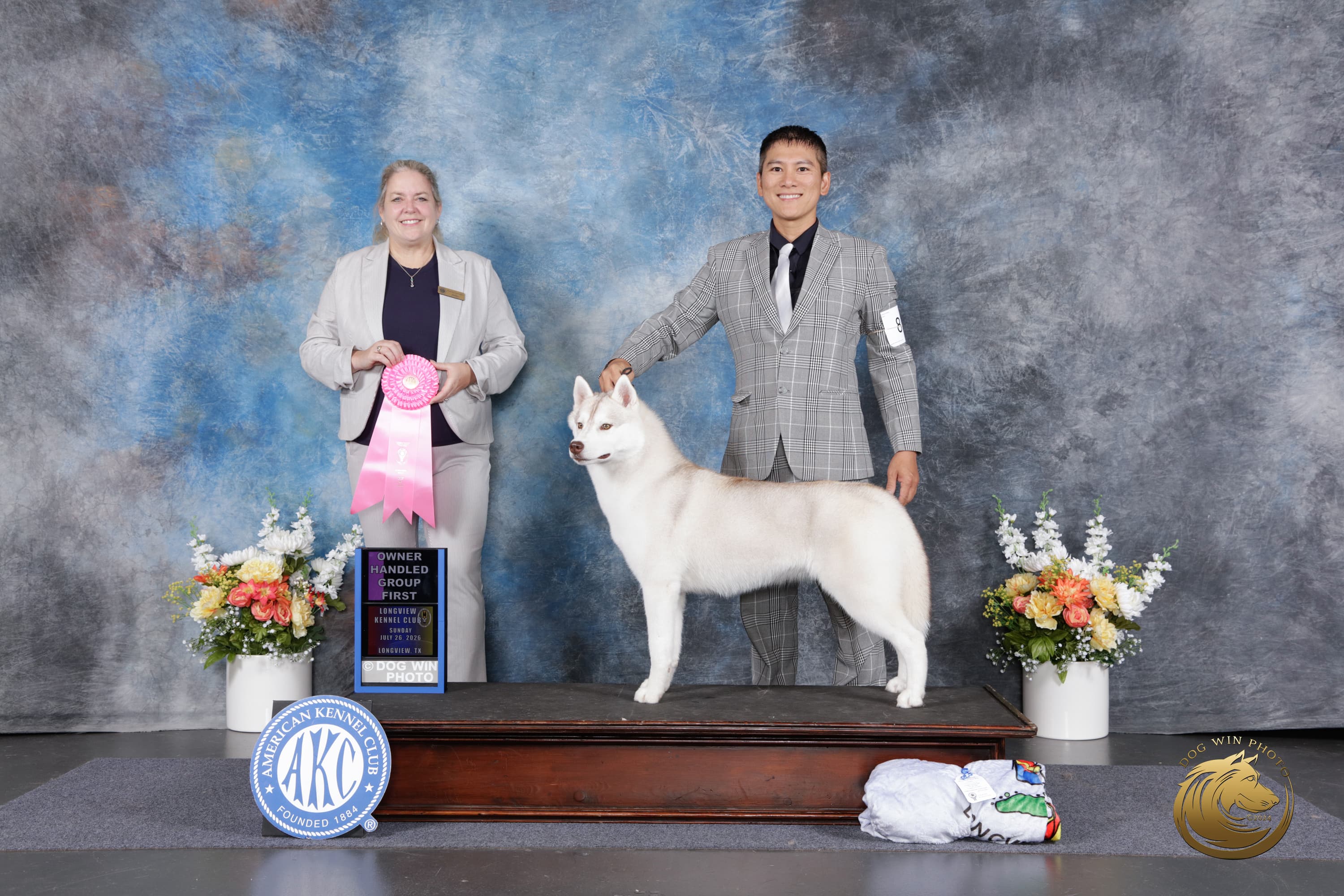 Caramel posed with her handler and Judge Pamela Schroeder beside a pink Owner-Handled Group First ribbon at Longview Kennel Club in 2026
