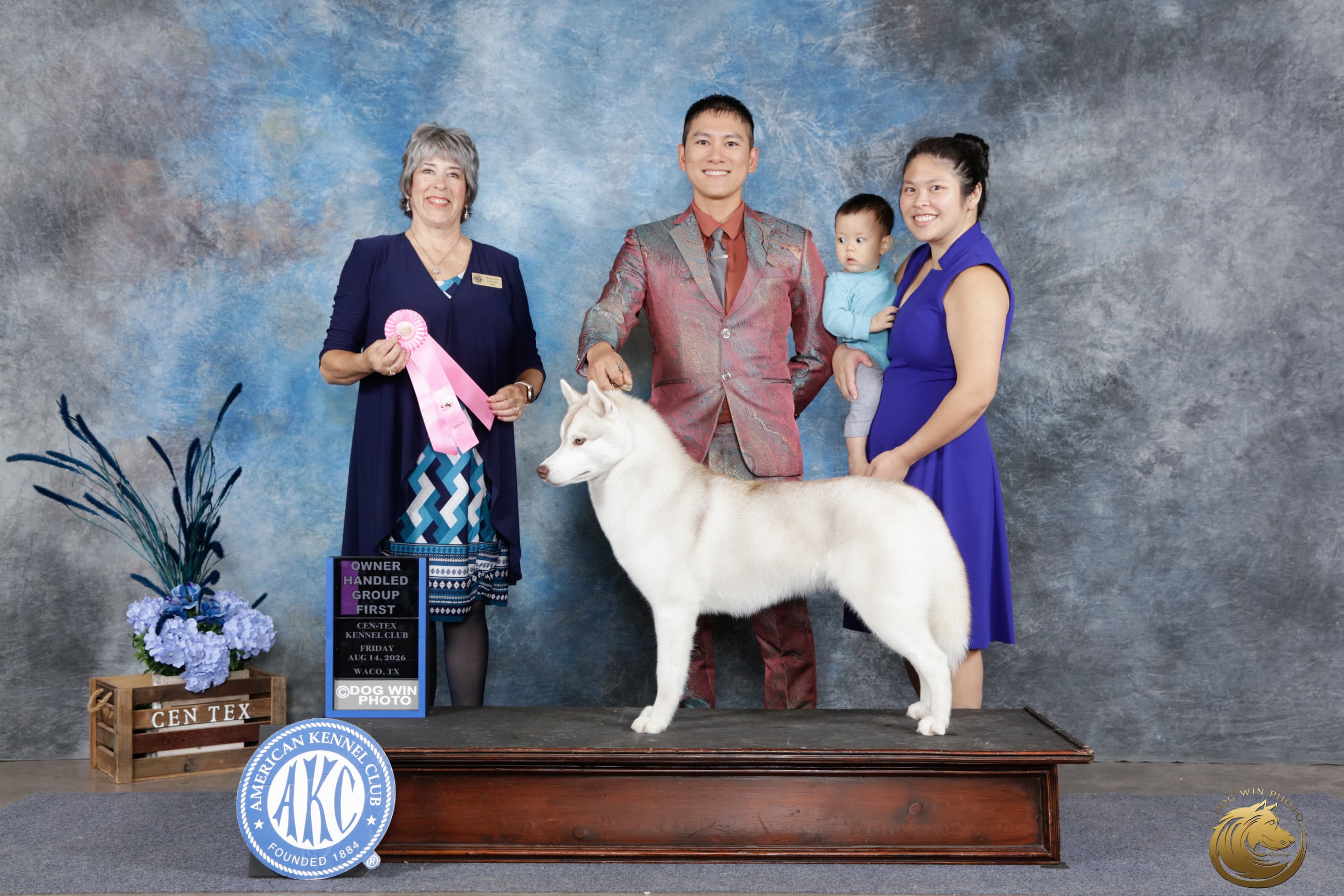 Caramel posed with her handler, family, and Judge Marie Falconer beside a pink Owner-Handled Group First ribbon at Cen-Tex Kennel Club in Waco in 2026